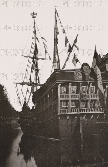 Galley on the Herengracht., Replica of a galley on the Herengracht during the April Festivals in 1887 in Amsterdam, photograph, cabinet photograph, Part of Photo album with images of the April Festivals in 1887 in Amsterdam., Amsterdam, 1887-04, photographer, pasteboard, ink, materials by function, paper, printing ink, collotype, photogravure, height 147 mm x width 97 mm, sailing-ship, sailing-boat, canal, prospect of city, town panorama, silhouette of city, April Festivals 1887, Herengracht (Amsterdam), A large ornate sailing ship is shown from the stern, with tall masts and rigging decorated with multiple triangular and rectangular flags; the ship’s stern has a multi-windowed gallery with curtained panes and decorative railings, and a carved emblem above the gallery. The hull is dark and weathered, and the ship is moored alongside a narrow waterway that reflects the vessel and nearby trees; portions of a canal bank and tree-lined shore are visible along one side. The sky is pale and ...