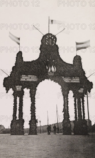 Nassauplein., Triumphal arch at Nassauplein during the April Festivals in 1887 in Amsterdam, photograph, cabinet photograph, Part of Photo album with images of the April Festivals in 1887 in Amsterdam., Amsterdam, 1887-04, photographer, pasteboard, ink, materials by function, paper, printing ink, collotype, photogravure, height 155 mm x width 95 mm, prospect of city, town panorama, silhouette of city, gate, entrance, April Festivals 1887, Nassauplein, A tall ornamental archway covered in dense foliage with multiple vertical pillars and rounded openings stands in the foreground, topped by a central rounded mound with small decorative elements and several flagpoles bearing flags; silhouettes of a few people are visible walking beneath the arch, and a row of thin masts and lamp posts can be seen in the background along a flat open area.