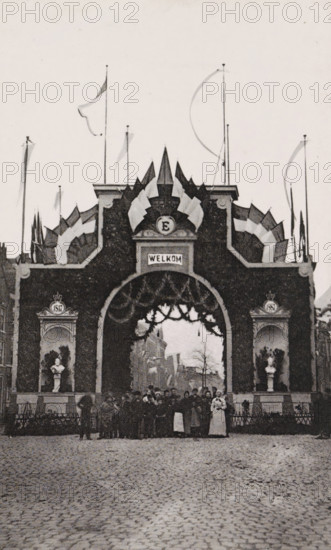 Westerstraat., Triumphal arch in the Westerstraat during the April Festivals in 1887 in Amsterdam, photograph, cabinet photograph, Part of Photo album with images of the April Festivals in 1887 in Amsterdam., Amsterdam, 1887-04, photographer, pasteboard, ink, materials by function, paper, printing ink, collotype, photogravure, height 147 mm x width 100 mm, prospect of city, town panorama, silhouette of city, gate, entrance, April Festivals 1887, Westerstraat (Amsterdam), A large ornate archway decorated with garlands and surrounded by stylized triangular pennants fills the center of the image, topped with tall poles bearing long flowing flags and streamers; two alcoves flank the arch, each containing a bust on a pedestal and framed by decorative molding, while a crowd of people stands beneath and in front of the arch on a cobbled street, with buildings and bare trees visible through the opening in the background.