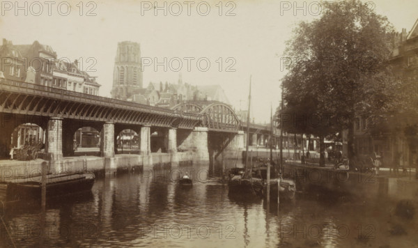 De Kolk. Rotterdam., View of De Kolk in Rotterdam with railway viaduct, photograph, cabinet photograph, Part of a Leporello album with photos of Rotterdam., photographer: attributed to Andries Jager, Rotterdam, c. 1860 - c. 1900, Attributed to, paper, pasteboard, albumen print, height 95 mm x width 156 mm, railway, train, bridge in village across river, canal, parts of church exterior and annexes: tower, viaduct, A sepia-toned riverside scene showing a long arched bridge with stone piers spanning across calm water, small boats moored and floating beneath and alongside the bridge, masts and rigging rising from some vessels, a quay with a low stone wall and wooden posts, a row of multi-story buildings with varied rooflines and dormer windows lining the far bank, a tall church tower and other roof silhouettes visible behind the buildings, a large leafy tree beside the water on the right bank, reflections of the bridge, boats, and tree in the water, and a few indistinct figures and horse-d...