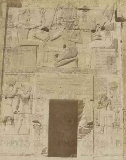 Wall with hieroglyphs and reliefs in Egypt (former title), Abydos Offrande de, Relief in the temple of Seti I at Abydos, photograph, Part of Travel album with 80 photos of Egypt., negative number: '883', photographer: Zangaki, C. & G., Egypt, c. 1870 - c. 1890, North Africa, pasteboard, paper, albumen print, height 276 mm x width 220 mm, Egyptian hieroglyphs, A stone relief panel showing carved figures arranged around a recessed dark doorway, with a central scene above the doorway of a kneeling figure facing a seated figure on the left and another seated figure on the right, each holding staffs and wearing ornate headdresses; the kneeling figure wears a pleated skirt and a tall, intricate headdress with curved elements and holds a curved staff; the seated figures wear tall crowns and collars and hold staffs and flails while seated on patterned thrones; below the central scene a horizontal band with a winged disk and carved panels extends above the doorway; flanking the doorway are vert...