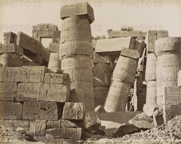 Columns with hieroglyphs of the Great Hall (Hypylical Hall) in the Temple of Karnak, Karnak Ruins of the Hypostyle Hall, interior view, Egypt, photograph, Part of Travel Album with 80 Photos of Egypt., negative number: '149', photographer: Maison Bonfils, Temple of Karnak, c. 1870 - c. 1898, North Africa, Luxor, pasteboard, paper, albumen print, height 221 mm x width 280 mm, temple, shrine, Egyptian religion, Egyptian hieroglyphs, A sepia-toned scene of ancient stone ruins with large carved columns and blocks arranged in a crowded, collapsed formation, showing detailed shallow relief carvings and repeating decorative motifs across weathered cylindrical and rectangular stone surfaces, some columns standing upright while others lean or lie broken among scattered rubble, a rectangular lintel visible near the top framed by stacked blocks, and a person wearing a head covering sitting on a stone ledge near the base of a leaning column.
