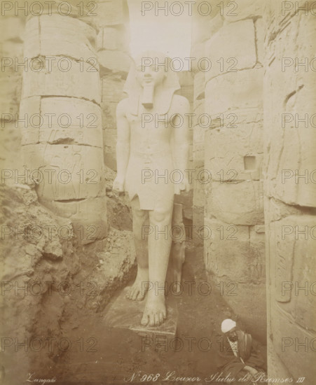 Statue of Pharaoh Ramses II in Luxor, Luxor Statue of Ramses III, photograph, Part of Travel Album with 80 Photos of Egypt., negative number: '968', photographer: Zangaki, C. & G., Luxor, c. 1870 - c. 1890, North Africa, pasteboard, paper, albumen print, height 276 mm x width 225 mm, Contrary to what the title on the object states, this is Ramses II, monument, statue, A large weathered stone statue of a standing pharaoh wearing a headdress and a false beard is centered between tall carved stone columns; the figure's arms hang at its sides and its legs and feet rest on a rectangular base, with visible carved details on the torso and kilt. Surrounding the statue are stacked stone blocks and columns with carved reliefs and worn surfaces, and rubble and loose stones at the base. In the lower right corner a seated man wearing a light head covering and layered clothing is partially visible, positioned near the statue's base and leaning against the stone wall. The overall scene has a muted, a...
