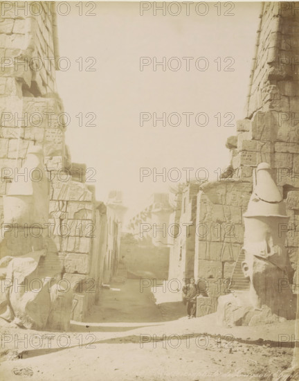 Porte triomphale de Louqsor (Egypt), Temple of Luxor, Colonnade and sculptures in Luxor (former title), photograph, Part of Travel album with 80 photos of Egypt., number: '41', negative number: '129', name: 'Bonfils, F. Bonfils', photographer: Félix Bonfils, Luxor, 1898, North Africa, pasteboard, paper, albumen print, height 279 mm x width 220 mm, piece of sculpture, reproduction of a piece of sculpture, gate, entrance, A faded sepia photograph shows a narrow ruined avenue flanked by tall stone walls and columns carved with reliefs, with large broken statues and fragments of sculpted heads and torsos leaning against the masonry on both sides; the stone surfaces display carved figures and decorative panels, and a set of column capitals is visible receding into the distance, while a solitary person in dark clothing stands near one of the right-hand statues, partially in shadow, and the foreground is a sandy, uneven ground leading into the sunlit passage.