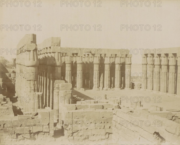 Louqsor. Ensemble de colonnade, Colonnade in Luxor, photograph, Part of Travel album with 80 photos of Egypt., negative number: '980', photographer: Zangaki, C. & G., Luxor, c. 1870 - c. 1890, North Africa, pasteboard, paper, albumen print, height 220 mm x width 276 mm, colonnade, A sepia-toned photograph of an ancient stone structure composed of rows of large vertical columns topped by rectangular beams, with some columns showing blocky capitals and horizontal stone lintels; the stone surfaces display weathering and chips, and scattered carved blocks and rubble lie in the foreground and between the columned aisles; shadows fall along the columns creating vertical dark bands, and a low horizon and pale sky fill the upper part of the image.