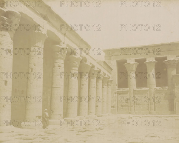 Courtyard of the Temple of Horus in Edfu, Edfou Coure de temple de Horous, photograph, Part of Travel album with 80 photos of Egypt., negative number: '...', photographer: Zangaki, C. & G., Temple of Horus (Edfu), c. 1870 - c. 1890, North Africa, pasteboard, paper, albumen print, height 217 mm x width 275 mm, temple, shrine, Egyptian religion, A row of tall stone columns with carved capitals lines a weathered temple facade, their surfaces marked with reliefs and horizontal grooves; adjacent to them is a second colonnade with similar capitals and rectangular openings above, and low stone walls and carved panels occupy the spaces between; the ground in front is uneven stone, and a person wearing light head covering and flowing garments stands near one of the columns.