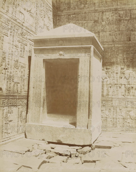 Interior of the Temple of Horus in Edfu, Edfu Cella de grand dans le temple de, photograph, Part of Travel album with 80 photos of Egypt., negative number: '1045', photographer: Zangaki, C. & G., Temple of Horus (Edfu), c. 1870 - c. 1890, North Africa, pasteboard, paper, albumen print, height 277 mm x width 221 mm, temple, shrine, Egyptian religion, Edfu, A stone shrine with a recessed rectangular opening stands in front of a wall covered in carved reliefs, the shrine featuring a sloped top and carved decorative band above the opening, flanked by vertical carved panels; the surrounding wall shows rows of figures and hieroglyphic-style carvings, and the stone floor in front of the shrine is uneven with scattered broken stones and slabs.
