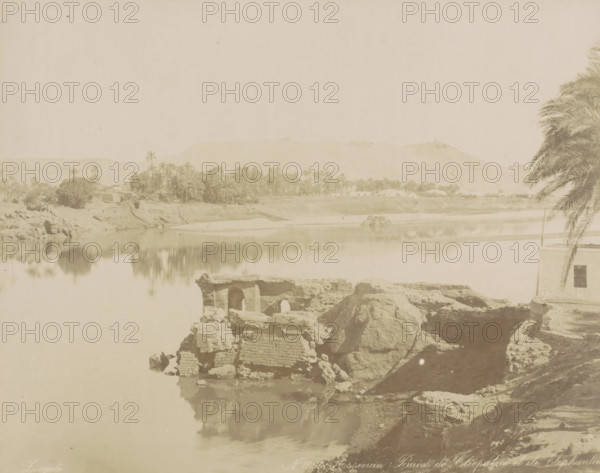 Assouan Bains de Cleopatra et ile Elephantine, Bath of Cleoptra and the island of Elephantine near Aswan, photograph, Part of Travel album with 80 photos of Egypt., negative number: '1060', photographer: Zangaki, C. & G., Aswan, c. 1870 - c. 1890, North Africa, pasteboard, paper, albumen print, height 218 mm x width 276 mm, island, river, Elephantine, A pale riverside scene showing a calm body of water reflecting the opposite bank, with clusters of palm trees and low buildings along the far shore, a range of low hills in the background, rocky ruins and large boulders in the foreground by the water's edge, and a partial view of a building with a window and an overhanging palm frond on the right side.