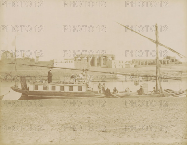Sailboat (dahabie) on the Nile with the Great Colonnade in Luxor in the background, photograph, Part of Travel album with 80 photos of Egypt., negative number: '126', photographer: attributed to Zangaki, C. & G., Luxor, photographer: attributed to Maison Bonfils, c. 1870 - c. 1890, Attributed to, pasteboard, paper, albumen print, height 220 mm x width 283 mm, Photos in this album are by the photographers Bonfils or by C. & G. Zangaki. Therefore, the photo has been attributed to them., sailing-ship, sailing-boat, river, temple, shrine, Egyptian religion, North Africa, Nile, A sepia-toned riverside scene showing a long wooden boat with a cabin and shutters along its side, a tall mast and horizontal spars, and several people on and around the boat wearing loose garments and head coverings; in the background across the water there are large stone buildings with columns and flat roofs and a low shoreline, and in the foreground a sandy, textured riverbank occupies the lower part of the image...