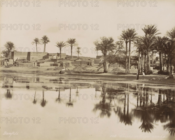Village with palm trees by a body of water near Cairo, Caire, village arabe, photograph, Part of Travel Album with 80 Photos of Egypt., negative number: '118', photographer: Maison Bonfils, Cairo (city), c. 1870 - c. 1898, Ottoman Empire, North Africa, pasteboard, paper, albumen print, height 223 mm x width 280 mm, prospect of village, silhouette of village, trees: palm-tree, A wide sepia-toned riverside scene shows a shoreline with mud-brick buildings and low ruined structures partially obscured by clumps of vegetation. Tall palm trees rise along the bank, some clustered and some standing alone, their fronds splayed at the tops. A pair of figures in long robes stand near the trees by the water's edge. Reflections of the trees and shoreline appear in the calm river in the foreground, creating mirrored shapes and soft ripples. The sky above is pale and empty, occupying the upper portion of the image.