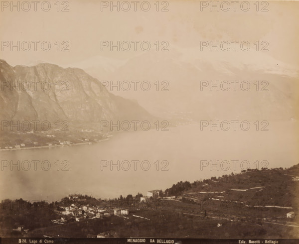 View of Menaggio from Bellagio, album page, photograph, This photo is part of an album., inscription: 'B28. Lado di Como, Menaggio da Bellagio, Ediz. Bosetti - Bellagio', annotation: '1898', photographer: Bosetti (possibly), Gotthard Pass, Bellagio (Italy), 1898, Lombardy, paper, albumen print, height 265 mm, width 350 mm, city-view in general, 'veduta', lake, mountains, Lake Como, Como, A sepia-toned panoramic view showing a large calm lake bordered by steep rocky mountains on one side and a gently sloping shoreline with scattered buildings and clusters of trees on the other, with a small village of tightly grouped houses in the foreground, terraced hillsides and winding roads, distant hazy peaks rising behind the nearer mountains, and a broad expanse of water occupying the central area of the image.