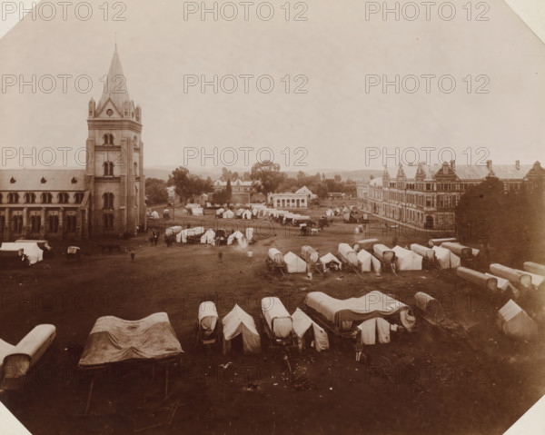 Church Square, Church Square with military encampment in Pretoria, photograph, Part of Album with 191 photographs of a trip to and through South Africa., Pretoria, c. 1885 - c. 1910, photographer, paper, daylight collodion silver print, height 234 mm x width 293 mm, (military) camp with tents, A wide sepia-toned scene shows a large open field filled with rows of covered wagons and canvas tents, with several small groups of people and horses scattered among them; on the left a tall stone building with a pointed spire and arched windows rises beside a long nave, and on the right a multi-story brick building with decorative gables and many windows frames the edge of the field; trees and smaller buildings sit beyond the tents, and low hills are visible along the distant horizon under a pale sky.