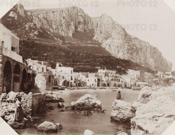 Island Capri, Capri la Marina Grande, View of the Marina Grande of Capri, photograph, Part of Album with 191 photos of a trip to and through South Africa., negative number: '2004', Capri, Isola di, publisher: Edizione Esposito, c. 1880 - c. 1920, photographer, paper, albumen print, height 198 mm x width 259 mm, harbour, A sepia-toned coastal scene showing a rocky shoreline and calm water in the foreground with large irregular rocks scattered along the shore and a person sitting on one rock facing the water. A woman stands on a rock near the left foreground wearing a long garment. Along the middle ground there is a row of arched stone structures and whitewashed buildings with balconies and doorways clustered at the water’s edge, and several small boats pulled up on the shore. Behind the buildings a terraced hillside rises with vegetation, and steep pale cliffs dominate the background, forming a high rocky mountain face that spans the image. The sky above is light and largely empty.