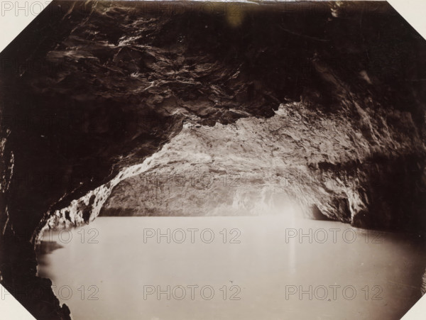 Capri la grotta azzurra preso dal vero, Interior of the Blue Grotto on Capri, Blue Grotto Capri, photograph, Part of Album with 191 photos of a trip to and through South Africa., negative number: '2094', Capri, Isola di, publisher: Edizione Esposito, c. 1880 - c. 1920, photographer, paper, albumen print, height 198 mm x width 258 mm, cave, grotto, A dark, irregular rocky ceiling with textured surfaces fills the upper portion of the image, transitioning to a lighter, rough rock surface toward the center where uneven striations and small protrusions are visible; the lower area shows a smooth, pale expanse with subtle reflections and faint speckles, and darker rock formations frame the left and right edges creating an archlike opening above the lighter central area.