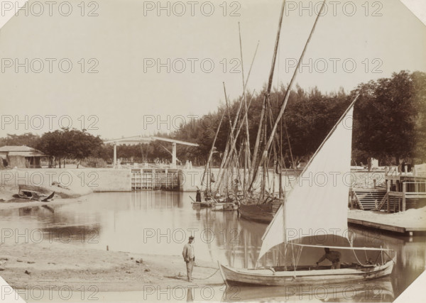 Sailing ships at a Suez Canal lock, Suez Canal, photograph, Part of Album with 191 photos of a journey to and through South Africa., c. 1870 - c. 1890, photographer, North Africa, paper, albumen print, height 195 mm x width 280 mm, canal, sluice, weir (landscape with figures, staffage), A calm river scene showing several sailboats moored along a bank, their tall masts clustered together and reflected in the water, a large triangular sail in the foreground partially obscuring a boat where a person leans over the gunwale, a second person standing on the sandy shore holding a rope, a small beached boat resting on the far bank, a wooden lock or gate structure spanning the water with a simple overhead mechanism, a cluster of trees and rustic buildings behind the bank, and wooden steps and a small pier area adjacent to the boats.