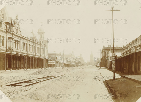 Pretoria, Broken-up Kirk Street with railway line in Pretoria, photograph, Part of Album with 191 photos of a trip to and through South Africa., c. 1890 - c. 1920, photographer, paper, daylight gelatin silver print, height 211 mm x width 292 mm, street (city(-scape) with figures, staffage), A wide dusty street flanked by long two-story buildings with covered arcades and decorative facades stretches toward a distant church tower, with an unpaved roadway and visible tram tracks in the foreground, a tall telegraph pole rising on the right, several smaller poles and signs along the sidewalks, ornate rooflines and a domed turret on the left, people and horse-drawn vehicles faintly visible far down the street, and piles of dirt and scattered stones along the road edges.