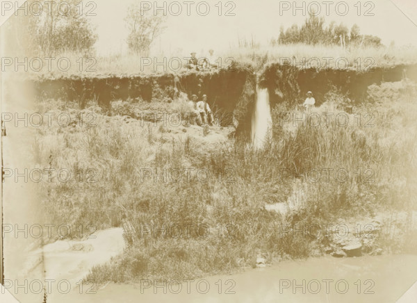 Near Pretoria, Europeans with a dog next to a waterfall near Pretoria, photograph, Part of Album with 191 photographs of a trip to and through South Africa., c. 1890 - c. 1920, photographer, paper, daylight gelatin silver print, height 213 mm x width 293 mm, waterfall, Pretoria, A pale sepia photograph showing a grassy riverbank with a vertical eroded cliff face and a narrow waterfall flowing from the top to the lower vegetation, tall grasses and reeds growing along the bank, scattered bushes and small trees behind the cliff, and five seated people wearing hats and light clothing positioned along the top and middle of the bank, with one person sitting alone near the right edge and others grouped nearer the center, all surrounded by natural terrain and vegetation.
