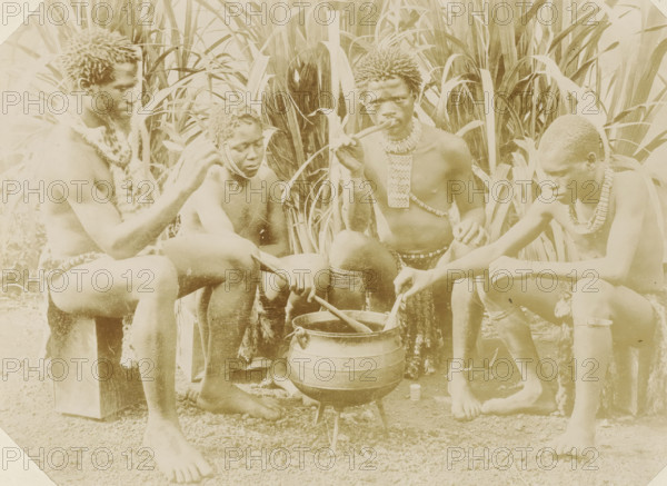 Four black men sitting around a cooking pot in South Africa, Kaffirs, photograph, Part of Album with 191 photographs of a trip to and through South Africa., South Africa, c. 1890 - c. 1920, photographer, paper, daylight gelatin silver print, height 213 mm x width 292 mm, Africans, kitchen-utensils: pot, Four shirtless people sit close together in front of tall, broad-leafed plants surrounding them. They all have short, textured hair and wear decorative necklaces and waist adornments made from beads and natural materials. One person on the left wears a fur-like garment draped over a shoulder and sits on a block while looking toward the center. Next to them, a person with beaded strands near their face looks downward while holding a stirring stick. In the center, a person holds a long pipe to their mouth and has layered necklaces and bracelets, with one hand resting on a knee. On the right, a person leans forward concentrating on stirring a large pot set on a small tripod over the ground...