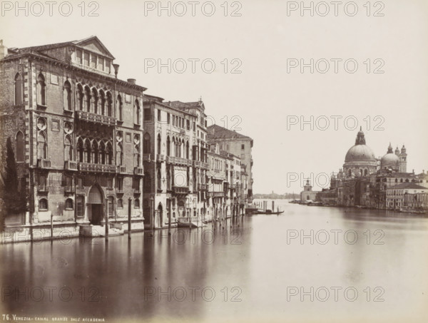 Venezia - Grand Canal from the Accademia, Grand Canal., Grand Canal with palaces, the Basilica of Santa Maria della Salute and the Fondamente Salute in Venice, photograph, Part of Topographical Album of Venice 1881., negative number: '76', check stamp: 'Carlo Ponti Venezia', photographer: Carlo Ponti, Venice, 1860 - 1881, paper, albumen print, height 200 mm x width 260 mm, canal, Roman Catholic Church, travel, Basilica of Santa Maria della Salute, Grand Canal, A riverside scene showing a wide waterway bordered by a row of ornate, multi-story brick and stone buildings with arched windows, balconies, decorative facades, and visible shutters on the left, wooden poles rising from the water along the building fronts, small boats moored at the edges, and a distant waterfront skyline on the right dominated by a large domed church with smaller domes and bell towers, all rendered in a monochrome photographic tone.