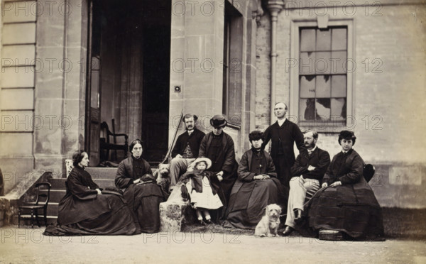 Portrait of the Peel family in front of a country house in Newbury, The Peel Family Newbury Somerset, photograph, Part of an English family album with photos of people, travel, cricket and artwork., Newbury (Somerset), c. 1860 - c. 1865, photographer, Somerset, paper, albumen print, height 125 mm x width 203 mm, Person, Peel, family, villa, dog, head-gear, dress, gown, trousers, breeches, clothing for the upper part of the body, three generations (family group), A group of people seated and standing on stone steps and in front of a building entrance, all wearing dark period clothing and hats, with one child in a light dress and bonnet seated at center, two small dogs near the front, a man standing behind the seated group, women with long skirts and shawls, a row of stone and brick architectural details including a doorway with an open interior and a tall window with broken panes, a chair visible inside the doorway, and a stone ledge and ground in the foreground.