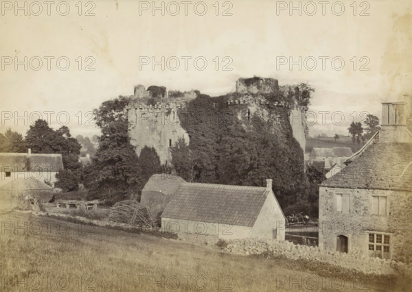 Nunney Castle Somerset, View of Nunney Castle and the surrounding buildings, photograph, Part of an English family album with photos of people, travel, cricket and artwork., Nunney, c. 1860 - c. 1870, photographer, Somerset, paper, albumen print, height 158 mm x width 208 mm, castle, ruin of a dwelling, house, plants and herbs: ivy, A sepia-toned scene showing a stone ruin partially covered in dense foliage rising behind a group of buildings, the ruin featuring battlemented tops and a round tower section. In front of the ruin, there are trees and hedges interspersed among a cluster of houses with pitched roofs, one roof tiled and another thatched, and a stone house with visible windows and a doorway. In the foreground a sloping grassy field leads down toward a low stone wall and scattered farm materials, including bundled branches and a wooden cart wheel. The sky above is pale and expansive, occupying the upper portion of the image.