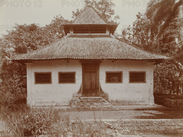 Missigit at Angké. No longer in use., Exterior of a mosque at Angké (Batavia), album page, photograph, Part of Travel Album with photos of Batavia., Batavia, c. 1920, photographer, Java, West Java, paper, daylight gelatin silver print, height 248 mm x width 305 mm, temple, shrine, Islam, Mohammedanism, overseas history, colonial history, Dutch East Indies, The, A single-story building with a tiled multi-tiered roof and a small upper pavilion sits centered in the image, flanked by dense trees and tall palm fronds on both sides; the building facade shows a central wooden door accessed by worn stone steps and framed by decorative trim, with two barred windows symmetrically placed on either side set into a smooth light-colored wall, and small exterior light fixtures under the roof eaves; the foreground contains overgrown grasses and scattered plant debris leading to a paved area in front of the steps.