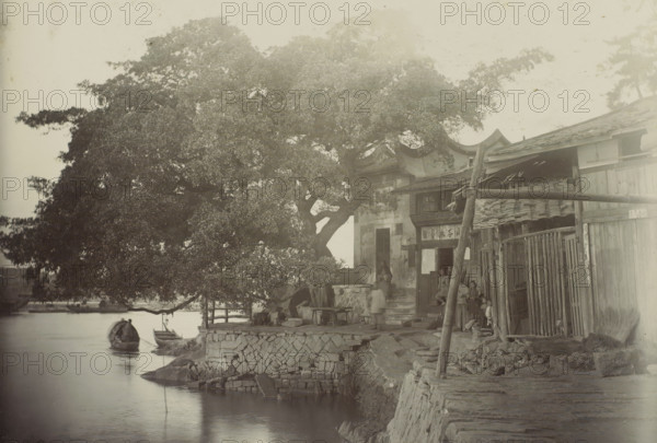 View of a house with a yard along the water, presumably in Fuzhou, photograph, Part of travel album by H. Dunlop with photographs of sights in Persia, Scotland, Germany, Russia, China, and Canada., photographer: Hendrik Dunlop (possibly), Fuzhou, c. 1890 - c. 1895, Fujian, paper, daylight gelatin silver print, height 106 mm x width 153 mm, courtyard, pier, quay, wharf, A riverside scene with a large leafy tree dominating the left and center, its branches extending over the water and a stone embankment. Small wooden boats float near the shore beneath the tree. Along the right side there are wooden buildings with tiled roofs and open doorways, a narrow stone path and steps leading up to one entrance. Several people are visible near the doorways and on the steps, some seated and some standing, with indistinct clothing and faces. Stacked tiles, wooden posts, and bamboo or wooden fencing are arranged against the buildings. The water surface is calm and reflective, and stonework forms the re...