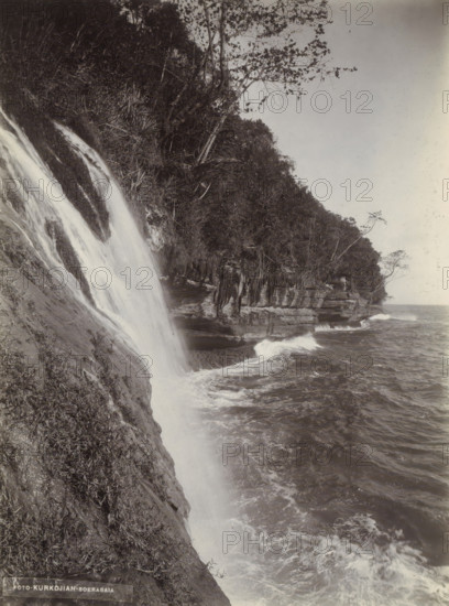 Coast of the Dutch East Indies with waterfall flowing into the sea, photograph, Part of Travel Album with forty photographs of the Dutch East Indies., photographer: Onnes Kurkdjian, Dutch East Indies, The, 1912, paper, daylight gelatin silver print, height 178 mm x width 237 mm, coast, waterfall, sea, A steep rocky shoreline with water cascading down a sloping rock face into a churning body of water, waves breaking against layered rock cliffs; dense vegetation and trees grow along the cliff edge, some branches extending outward, and the sky appears clear above the treeline.