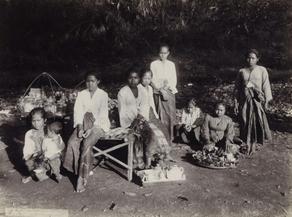 Women and children with goods displayed along the side of the road, Indies women and children with goods displayed along the side of the road (former title), photograph, Part of Travel Album with Forty Photos of the Dutch East Indies., photographer: Onnes Kurkdjian, Dutch East Indies, The, 1912, paper, daylight gelatin silver print, height 176 mm x width 238 mm, merchant, salesman, selling, A group of people arranged outdoors on bare ground in front of a backdrop of dense foliage, with several seated on low stools or the ground and a few standing; most wear patterned lower garments and light-colored tops, and some have scarves or shawls draped around their shoulders; one person holds a small child on their lap; in front of the group are small trays and containers that include jars, a teapot, cups, and other assorted vessels, and a woven tray with wrapped items; two curved supports rise from a low table that holds additional jars and small objects; the lighting creates strong contrasts ...