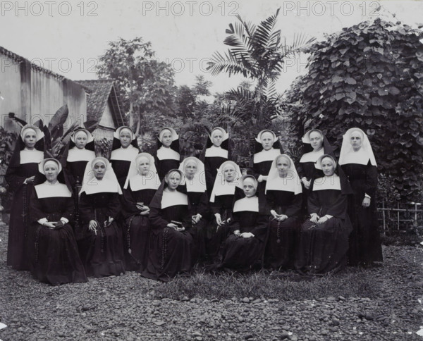 The Novices., Group portrait of the novices of the Ursulines, presumably in Weltevreden, album page, photograph, Part of Album with photos of Batavia and of the Ursuline sisters., Weltevreden, c. 1890 - c. 1910, photographer, Java, West Java, Batavia, paper, daylight collodion silver print, height 217 mm x width 270 mm, nun(s), A black and white photograph showing a group of women wearing traditional religious habits with white wimples and dark veils, arranged in rows outdoors; most are seated while others stand behind them, many holding rosary beads or small books in their hands, and several wear glasses. The background includes dense tropical vegetation with large leafy plants and a tall palm, a tiled roof and a wall partially visible to one side, and a gravel foreground with a small patch of grass near the front.