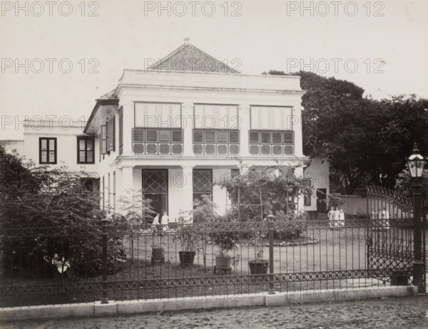Exterior of the Ursuline Convent of Noordwijk (Batavia), Convent of Noordwijk, front., album page, photograph, Part of Album with photos of Batavia and of the Ursuline sisters., Batavia, c. 1890 - c. 1910, photographer, Java, West-Java, paper, albumen print, height 216 mm x width 278 mm, abbey, monastery, convent, Roman Catholic church, façade (of house or building), A two-story colonial-style house with a tiled, pyramidal roof and a broad upper facade featuring large shuttered windows; the ground level has a covered porch supported by columns and doors with diamond-pane window panels; a wrought-iron fence and gate run across the front of the property with potted plants and small trees placed inside the yard; dense foliage and large trees flank the building on both sides; a tall street lamp stands outside the fence near the gate; several people in light clothing stand near the porch and by the gate.