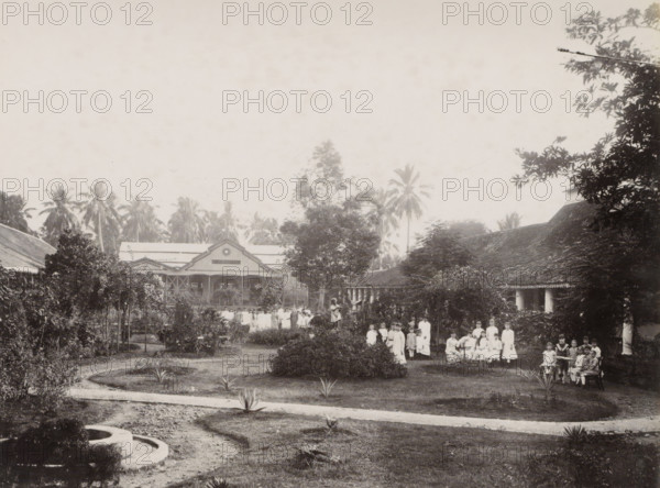The garden of the convent, Noordwijk., Girls in the convent garden of the Ursuline Sisters of Noordwijk (Batavia), with a building with the inscription 'Bloemendaal' in the background, album page, photograph, Part of Album with photos of Batavia and of the Ursuline Sisters., Batavia, c. 1890 - c. 1910, photographer, Java, West-Java, paper, albumen print, height 216 mm x width 279 mm, garden, abbey, monastery, convent, Roman Catholic church, child, A wide garden scene with a curved dirt path and several small spiky plants in the foreground leading toward a central grassy area with low shrubs and taller bushes, framed by trees and hedges on either side; a group of people, many in light-colored dresses and some seated, are gathered on the right side near a veranda and among the shrubs, with additional figures standing further back along a central walkway; behind them are two large buildings with pitched roofs and decorative gables partially obscured by trees, and tall palm trees rising be...