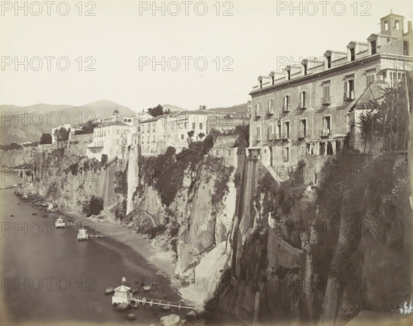 Sorrento!, Houses along a cliff in Sorrento, photograph, Part of Travel album with photos of sights in Naples and the surrounding area., Sorrento, c. 1865 - c. 1875, photographer, paper, albumen print, height 200 mm x width 251 mm, coastal cliff or crag, A seaside cliffside scene showing multiple buildings perched along the cliff edge, with a large multi-story building on the right featuring rows of windows and small balconies and a series of dormer windows on the roof, and a row of smaller light-colored buildings extending leftward along the cliff. Vegetation and trees appear among the buildings and on the cliff face, with steep rocky cliffs dropping to a narrow shoreline. Small pavilions on piers extend into the water below, connected by narrow walkways, and small boats are moored near the shore. Hills and distant mountains form the background under a pale sky.