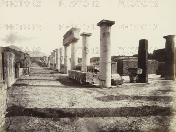 Pompeii forum: excavations 1813., Remains of buildings in Pompeii, photograph, Part of travel album with photos of sights in Naples and the surrounding area., negative number: '4200', Pompeii, c. 1865 - c. 1875, photographer, paper, albumen print, height 205 mm x width 252 mm, column, pillar, architecture, excavation, archaeology, A wide sunlit scene of ancient stone ruins with a long colonnaded walkway receding into the distance, showing rows of cylindrical stone columns and rectangular stone blocks aligned along the path. The foreground features a textured dirt and gravel surface with shadows cast by the columns, and several tall columns with square capitals stand prominently mid-frame. Additional broken stone elements and low walls with patterned brickwork appear to the right, while on the left are remnants of stone facades and niches opening onto the walkway. Low hills and a plume of smoke or cloud rise in the distant background beneath a pale sky.
