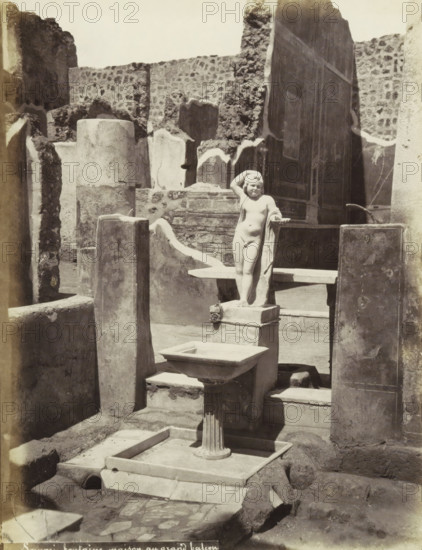 Pompeii fountain house with a large balcony, Remains of a house in Pompeii with a statue and a basin, photograph, Part of a travel album with photos of sights in Naples and the surrounding area., Pompeii, c. 1865 - c. 1875, photographer, paper, albumen print, height 256 mm x width 195 mm, excavation, archaeology, piece of sculpture, reproduction of a piece of sculpture (sculpture), A sunlit archaeological courtyard filled with weathered stone walls and broken pillars surrounds a small marble statue of a nude child standing on a pedestal, one hand raised to the head and the other resting on a draped cloth, with a basin and ornate columned fountain in the foreground and irregular stone paving and scattered fragments visible around the base.