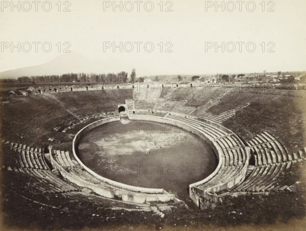 Interior of the amphitheater of Pompeii, Pompei: Amphitheatre. (pour 20 mille spectators) excavations 1813., photograph, Part of Travel Album with photos of sights in Naples and the surrounding area., negative number: '4209', Pompeii, c. 1865 - c. 1875, photographer, paper, albumen print, height 203 mm x width 252 mm, theater (building) - AA - open-air performances, A broad view of a circular amphitheater with concentric stone seating rising from a central flat arena, showing worn steps and curved retaining walls; a single small figure stands in the center of the arena; the seating tiers show gaps, diagonal stairways, and entry passages, one of which is a dark arched tunnel opening onto the arena; beyond the amphitheater, a low stone perimeter and a line of trees stretch across the horizon, with scattered buildings and distant vegetation under a pale sky.