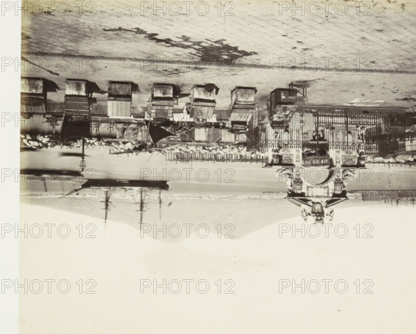 Harbor of Naples in the Santa Lucia neighborhood with oyster vendors' stalls in the foreground, S. Lucia. La Plage d'huîtres, photograph, Part of Travel Album with photos of sights in Naples and the surrounding area., photographer: attributed to Giorgio Sommer, Naples, c. 1865 - c. 1875, Attributed to, paper, albumen print, height 199 mm x width 250 mm, stall, booth (, market), harbor (view, vehicle, ship, etc.), An upside down black and white photograph showing a waterfront scene with a paved promenade, a row of wooden market stalls and carts, a wrought iron fence with decorative stone pillars and sculptural details, scattered crates and awnings, a lamppost, piles of objects near the water’s edge, reflections of masts and rigging in the water, and distant hills on the horizon.