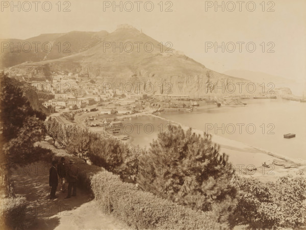 View of the port of Oran, Oran. Le Djabel Mourdjadjo et le Port, vue prize de la promenade de Letang, album page, photograph, This photo is part of an album., number: '204', photographer: Étienne Neurdein, c. 1880 - c. 1890, paper, albumen print, albumendruk, height 207 mm x width 272 mm, harbour, city-view in general, 'veduta', mountains, North Africa, Oran, A sepia-toned panoramic view showing a coastal town at the foot of a large rocky hill, with clustered buildings and docks along a calm bay, a breakwater extending into the water, small boats moored near the shore and one floating further out, steep cliffs rising behind the harbor, terraced slopes and scattered structures on the hillside, foreground vegetation including trees and hedges, and three men dressed in dark coats and hats standing on a shaded path overlooking the scene.
