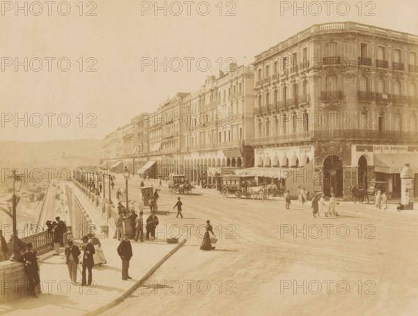 View of Boulevard de la Republique in Algiers, Alger. - Boulevard de la Republique, album page, photograph, This photo is part of an album., number: '284', photographer: Étienne Neurdein, Algiers, 1880 - 1890, North Africa, paper, albumen print, albumendruk, height 208 mm x width 273 mm, historical persons, avenue, boulevard, promenade, esplanade, A wide street scene with a long row of ornate multi-story buildings featuring balconies and arched ground-level openings along the right side, horse-drawn carriages and a larger horse-drawn omnibus on the roadway, groups of people walking and standing on the sidewalk and promenade, lampposts lining the elevated promenade with a balustrade, awnings and canopies over some shopfronts, and distant low hills visible in the background.