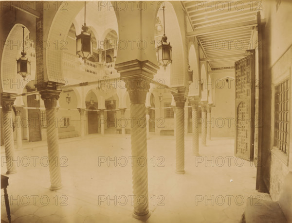 Courtyard of Palais des Raïs in Algiers, Alger. Interior du Palais du Gouverneur, album page, photograph, This photo is part of an album., number: '224', photographer: Étienne Neurdein, c. 1880 - c. 1900, paper, albumen print, albumendruk, height 210 mm x width 272 mm, arch, archivolt, architecture, column, pillar, palace, North Africa, Algeria, A vaulted interior space with tall arches and ornate carved columns featuring twisted shafts and decorated capitals, hanging lanterns suspended from the arches, an open carved wooden door on the right, rows of wooden beams across the ceiling at the right, a chandelier suspended in the central background, patterned wall panels and small balconies above, tiled floor, benches and doorways along the far walls, and decorative latticework on the right window.
