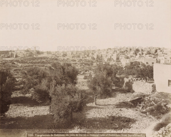 General view of Bethlehem from David's well. - General view of Bethlehem from David's well., View of Bethlehem from David's Well, photograph, Part of Travel Album with Photos of Jerusalem and Other Biblical Places., negative number: '1225', photographer: Maison Bonfils, Bethlehem (Israel), c. 1867 - c. 1895, Maison Bonfils, photographer, Palestine, pasteboard, paper, albumen print, height 224 mm x width 284 mm, prospect of village, silhouette of village, A wide panoramic scene showing a hillside landscape with scattered trees and terraced ground in the foreground and a dense cluster of flat-roofed stone buildings on a rise in the background, with a few larger structures that have towers or domes among the buildings; stone walls and paths are visible winding through the terrain, and vegetation appears sparse and scrubby across the slopes.