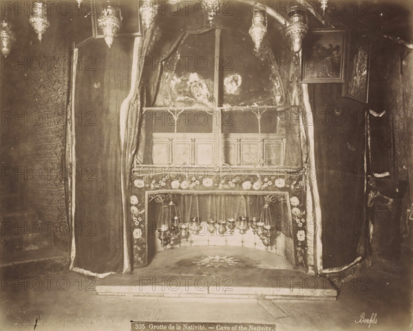 Grotte de la Nativite.- Cave of the Nativity., Nativity Cave in the Church of the Nativity in Bethlehem, photograph, Part of Travel Album with Photos of Jerusalem and Other Biblical Places., negative number: '335', photographer: Maison Bonfils, Bethlehem (Israel), c. 1867 - c. 1895, Palestine, pasteboard, paper, albumen print, height 225 mm x width 283 mm, interior of church, cave, grotto, A sepia-toned interior scene showing a small recessed alcove framed by heavy draped curtains with patterned borders and floral trim, a decorative metal grille across the upper front of the recess, hanging ornate lamps suspended inside the lower portion, a stone floor with a star-like design in front of the alcove, textured walls and framed pictures mounted on the wall to the right, and additional ornate hanging lamps visible along the top edge.