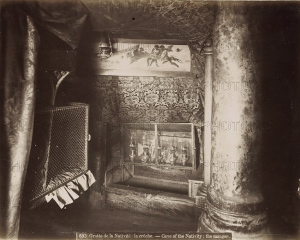 Crib in the Cave of the Nativity in Bethlehem, Grotte de la Nativite: la crèche.- Cave of the Nativity: the manger., photograph, Part of Travel Album with Photos of Jerusalem and Other Biblical Places., negative number: '893', photographer: Maison Bonfils, Bethlehem (Israel), c. 1867 - c. 1895, Palestine, pasteboard, paper, albumen print, height 222 mm x width 282 mm, cradle, crib, A dim interior scene with patterned fabric draped across the ceiling and walls, a hanging ornate lamp near the top center, a painted panel showing figures and clouds partially visible above the fabric, a decorative tapestry with intricate motifs covering the back wall, a metal mesh enclosure with objects behind it in the middle foreground, heavy textured columns on the right and left edges, and a ruffled cloth hanging from a mesh panel on the left.