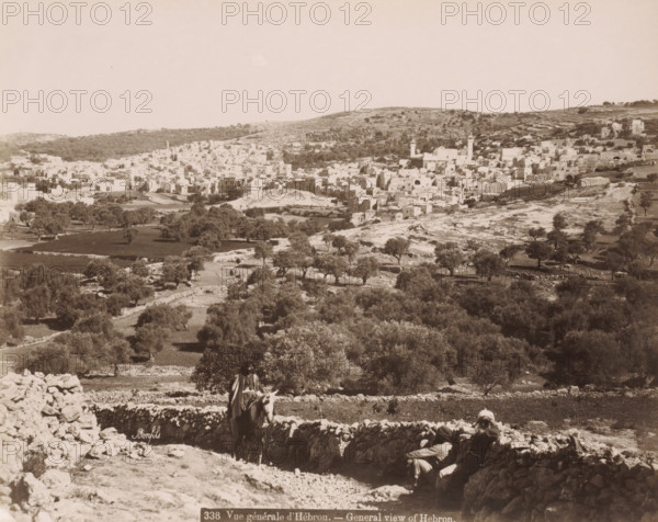 General view of Hebron.- General view of Hebron., View of Hebron, photograph, Part of Travel Album with photos of Jerusalem and other biblical places., negative number: '338', photographer: Maison Bonfils, Hebron, c. 1867 - c. 1895, Palestine, West Bank, pasteboard, paper, albumen print, height 222 mm x width 279 mm, prospect of city, town panorama, silhouette of city, A wide sepia-toned landscape showing a hillside town with densely packed light-colored stone buildings and a few taller tower-like structures set against rolling hills, with scattered trees and terraced fields in the foreground; a dry stone wall runs across the lower part of the scene, where two figures sit and one figure rides a donkey near the wall, and further down the slope there are more scattered trees and rocky ground.