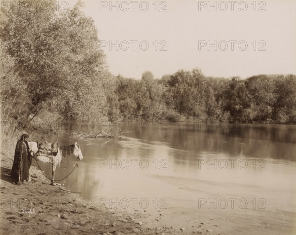 Cours du Jourdain.- The course of the Jordan, Man with a horse on the bank of the Jordan, photograph, Part of Travel album with photos of Jerusalem and other biblical places., negative number: '916', photographer: Maison Bonfils, Jordan (river), c. 1867 - c. 1895, pasteboard, paper, albumen print, height 227 mm x width 284 mm, riding a horse, ass, or mule, rider, horseman, river (landscape with figures, staffage), A riverside scene with calm reflective water edged by a tree-lined far bank and a large leafy tree on the near left, a person standing at the water's edge holding a rope attached to a saddled horse that faces the water, the person wearing a long dark garment and a head covering and leaning on a long staff, vegetation and low shrubs along the shoreline, and a sandy, pebbled foreground leading to the water.