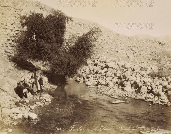 Men at the well Ein as-Sultan near Jericho, Fontaine d'Elisee. - Aïn-el-Sultan, près Jéricho, photograph, Part of Travel album with photos of Jerusalem and other biblical places., negative number: '345', photographer: Maison Bonfils, Jericho, c. 1867 - c. 1895, Maison Bonfils, photographer, Palestine, West Bank, pasteboard, paper, albumen print, height 223 mm x width 281 mm, well, A rocky riverside scene with a narrow stream flowing toward the foreground, smooth water reflecting light and rippling around scattered stones, a large leafy tree with dense foliage standing near the water's edge and casting a shadow over part of the stream, a hillside covered with loose rocks rising behind the tree and extending across the background, several people wearing layered garments and head coverings standing and sitting on the rocky bank near the tree, one person holding a long staff, and sparse low vegetation among the stones.