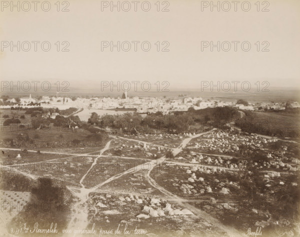View of Ramla in Israel, Ramallah and surrounding land, seen from a tower (former title), Rameleh, vue générale prise de la tour, photograph, View of Ramla with the Plain of Sharon and the Mountains of Judea, in the foreground an Islamic cemetery. Part of Travel Album with photographs of Jerusalem and other biblical places., negative number: '241bis', photographer: Maison Bonfils, Ramallah, c. 1867 - c. 1895, Palestine, pasteboard, paper, albumen print, height 225 mm x width 283 mm, prospect of city, town panorama, silhouette of city, Ramla, A sepia-toned aerial view shows a broad landscape with a foreground of rocky, uneven ground marked by winding dirt paths and scattered stone markers forming small rectangular shapes, some grouped and some isolated; patches of low vegetation and occasional trees dot the area. In the middle distance a belt of darker trees and denser foliage separates the foreground from a low-lying town of light-colored buildings with flat and pitched roofs, among wh...