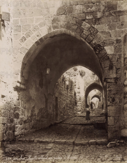 Jérusalem - Ruelle allant au palais d'Hérode, Street to Herod's Palace in Jerusalem, photograph, Part of Travel album with photos of Jerusalem and other biblical places., negative number: '259', photographer: Maison Bonfils, Jerusalem, c. 1867 - c. 1895, pasteboard, paper, albumen print, height 282 mm x width 222 mm, arch, archivolt, architecture, sloping street, street with steps or stairs, A narrow cobblestone passageway lined with rough stone walls and successive stone arches receding into the distance is shown, with light filtering through the arch openings and a hanging lantern on the left wall under the nearest arch; a person wearing a long garment stands facing away in the middle distance while another figure is farther along under the next arch, and the walls display patches of plaster, weathered stone blocks, and a small window grate high on the left side.