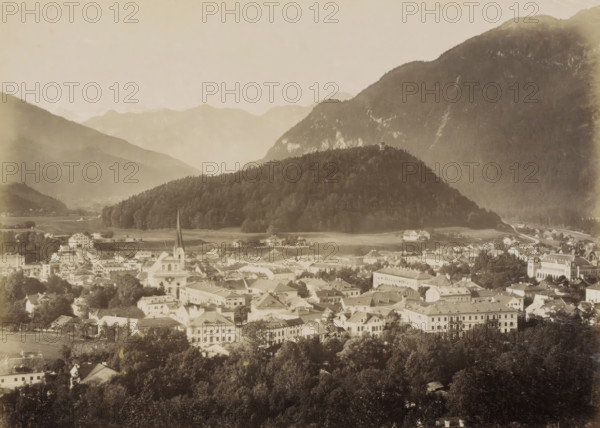 Ischl vom Gassner, View of Ischl and surrounding mountains from the Gassner, photograph, Part of travel album with photos of sights in Germany and Austria and of works of art., negative number: '345', Ischl, c. 1870 - c. 1900, photographer, Upper Austria, paper, albumen print, height 198 mm x width 267 mm, prospect of city, town panorama, silhouette of city, A wide sepia-toned view of a town nestled at the base of a densely wooded hill and surrounded by larger mountains, showing a foreground of clustered buildings with pitched roofs and varied architectural styles, interspersed with trees and open spaces, and a prominent church with a tall pointed spire rising above nearby structures, while the midground features a tree-covered hill topped by a small tower, and the background reveals layered mountain slopes fading into the sky.