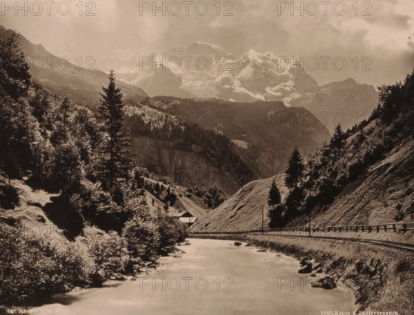 Route to Lauterbrunnen., Mountain landscape with the railway from Interlaken to Lauterbrunnen, album page, photograph, Part of Travel album with sights in Germany, Austria, Switzerland, Luxembourg and Belgium., number: '1023', photographer: Schroeder & Cie., c. 1880 - in or before 1899, ink, materials by function, paper, printing ink, collotype, photogravure, height 210 mm x width 275 mm, railway, train, mountain-range, snow peaks, A sepia-toned mountain scene with a river flowing toward the foreground, flanked by grassy slopes and rocky banks. Evergreen trees and clusters of leafy trees rise on both sides of the valley, with taller conifer silhouettes standing prominently against the sky. A narrow road or rail line runs along the right bank, bordered by a low fence and a few utility poles. A small building with a sloped roof sits near the base of a hillside in the midground. Layered ridges and forested hills lead back to a jagged, snow-covered mountain range under a cloudy sky.